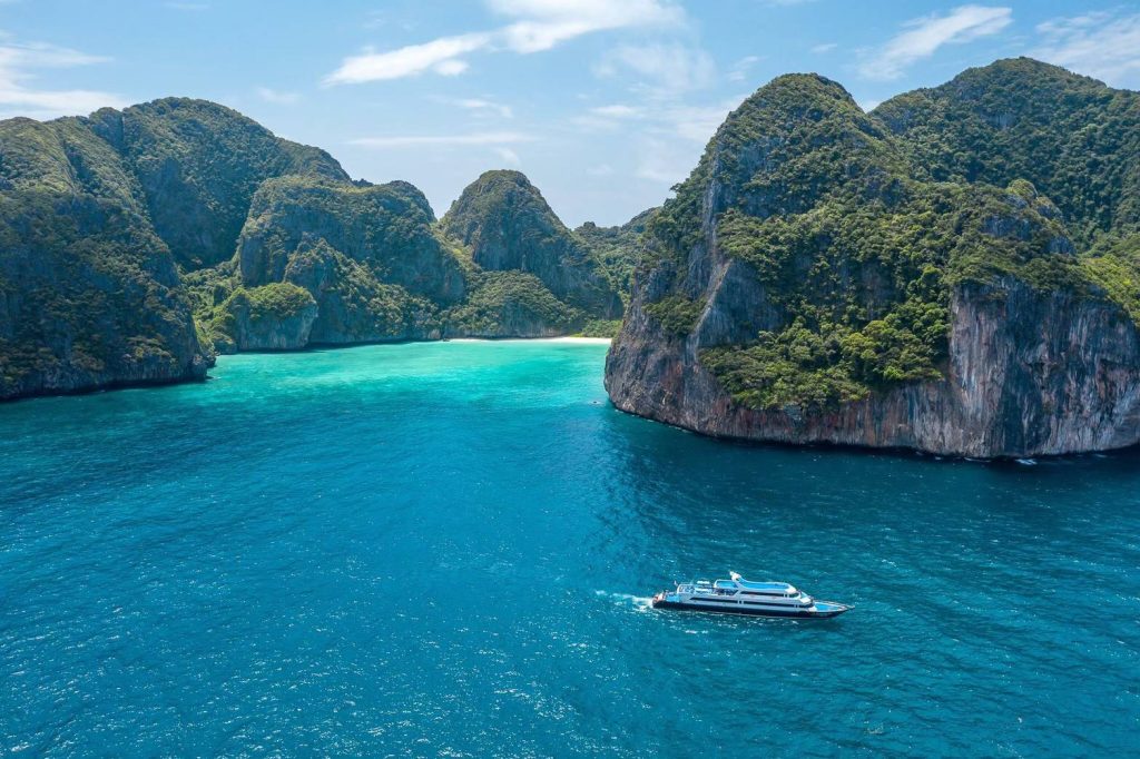 A scenic view of a luxury boat cruising through the crystal-clear turquoise waters near the limestone cliffs of Phi Phi Islands in Thailand. A top highlight of Thailand tours, this tropical paradise is known for its stunning beaches, vibrant marine life, and dramatic coastal landscapes.