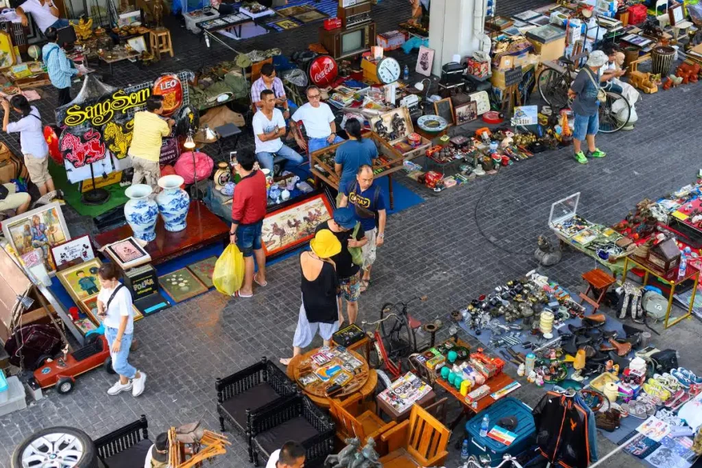 A vibrant flea market scene in Bangkok, Thailand, with various stalls selling antiques, vintage items, and collectibles. People are browsing through unique goods like old toys, ceramics, paintings, and clothing. A popular stop for Thailand tours, the market offers a glimpse into local culture and everyday life